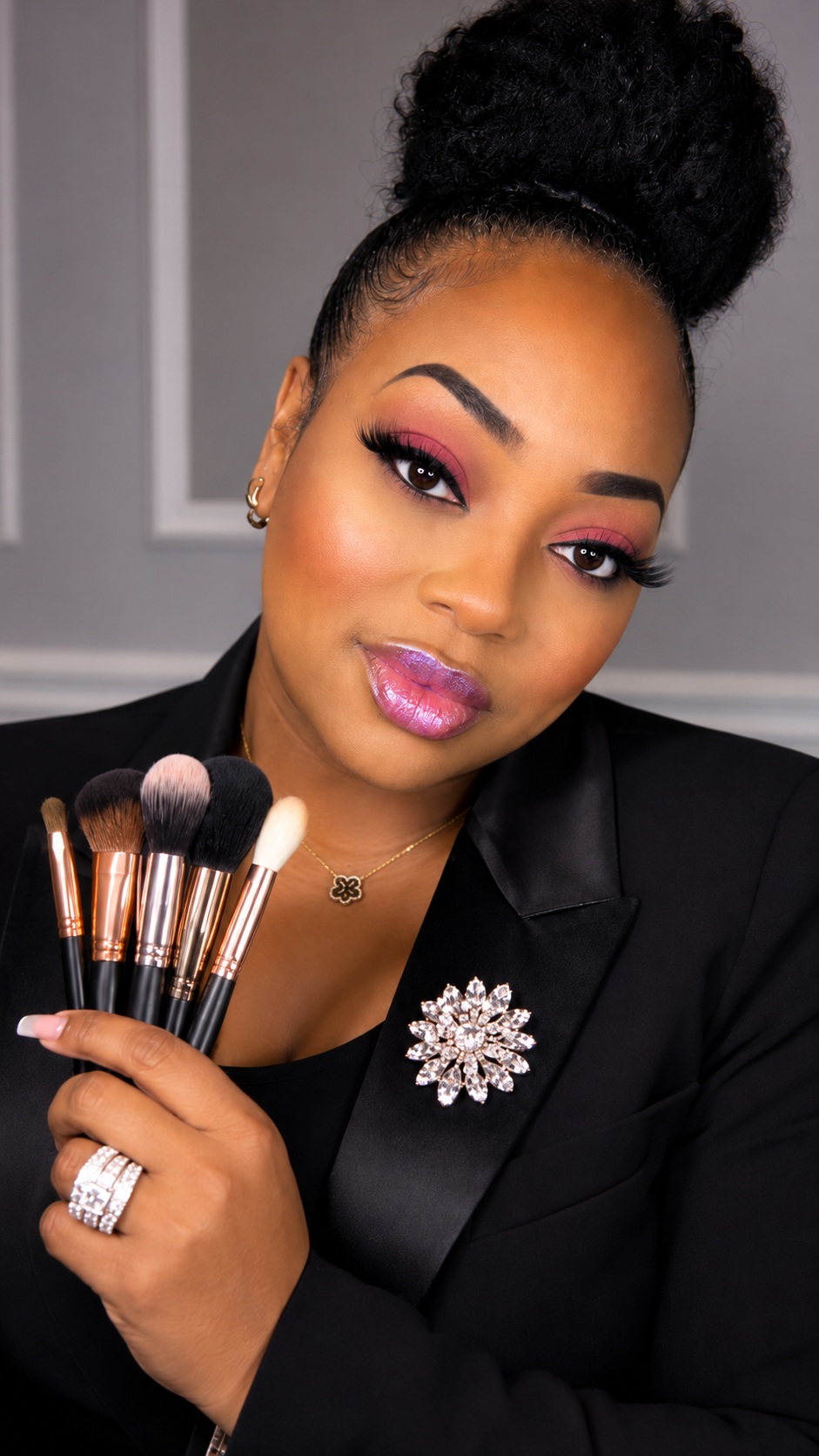 Matika Miss Tika Goldsmith holding makeup brushes, wearing a black blazer with a sparkling brooch